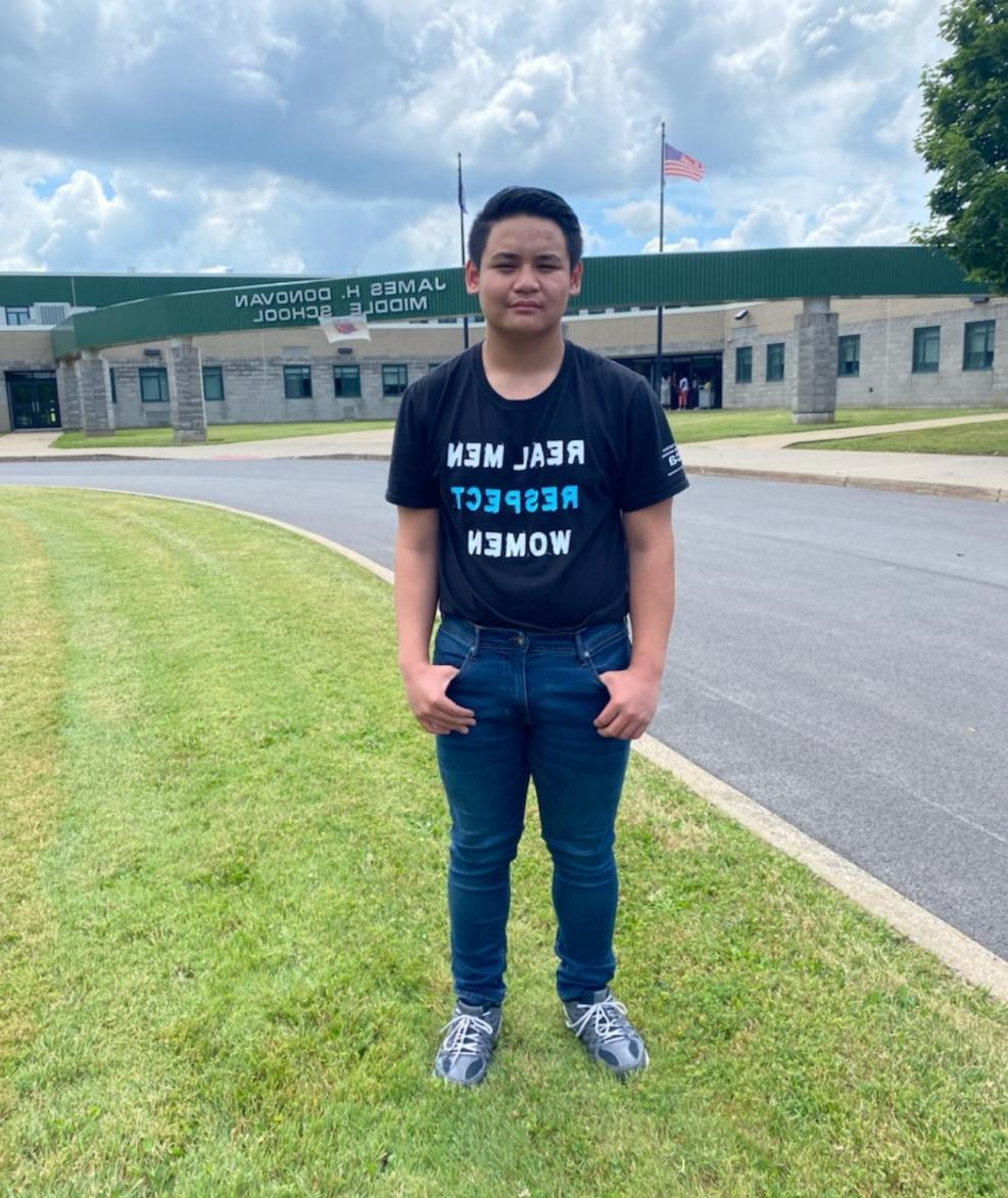 A young male student standing in front of Donovan Middle School wearing an 一起修改 shirt.